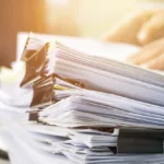 Stacks of white papers joined by binder clips with a hand lying on top of the pile