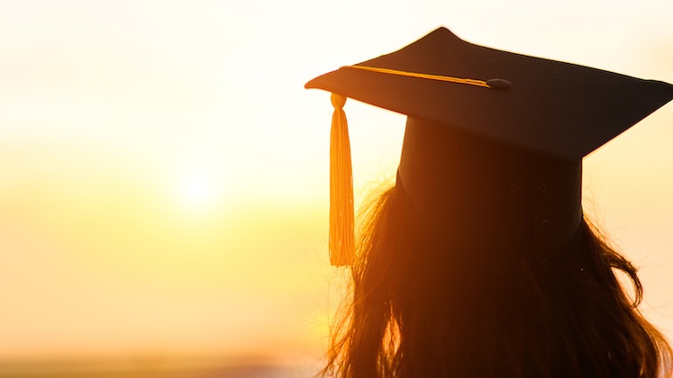Silhouette of a woman wearing a graduation cap staring into a setting sun.