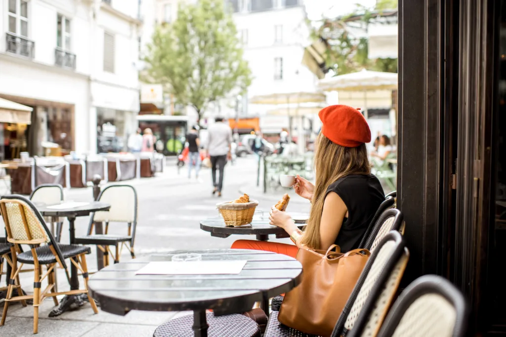 Woman sits at cafe in Paris wearing a red beret and eating a baguette.