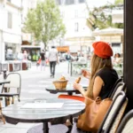 Woman sits at cafe in Paris wearing a red beret and eating a baguette.