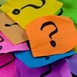 A pile of colorful sticky notes in lime, yellow, orange, pink, purple, and blue each have a thick black question mark written on them in marker.