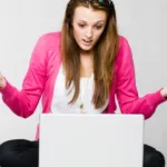 Young woman with long brown hair wearing a pink cardigan, a white shirt, and black pants sits in front of a laptop with her hands in the air looking despondent.