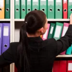 Person stands in front of a wall of binders of different colors with hands reaching for binders on different shelves.