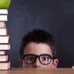 Face with short hair and glasses peers above a wooden table with stack of 9 books and an apple to the right of the face; copy tests are nothing to be scared about!