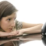 Woman lays head on her hands which sit on top of a desk as she stares at the phone in front of her.