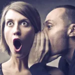 Man whispers into woman's ear and woman wears a shocked expression on face with open mouth and bulging eyes.