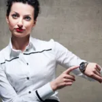 Brundette with hair tied back wearing red lipstick and a white, button-down shirt points to her wristwatch and looks fatigued