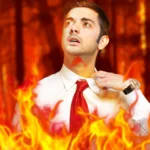 Man in white button down with red tie pulls on his collar to fan himself while flames engulf him.