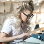 Blonde woman with curly hair wearing thick black glasses and a white, short-sleeved shirt types on a typewriter