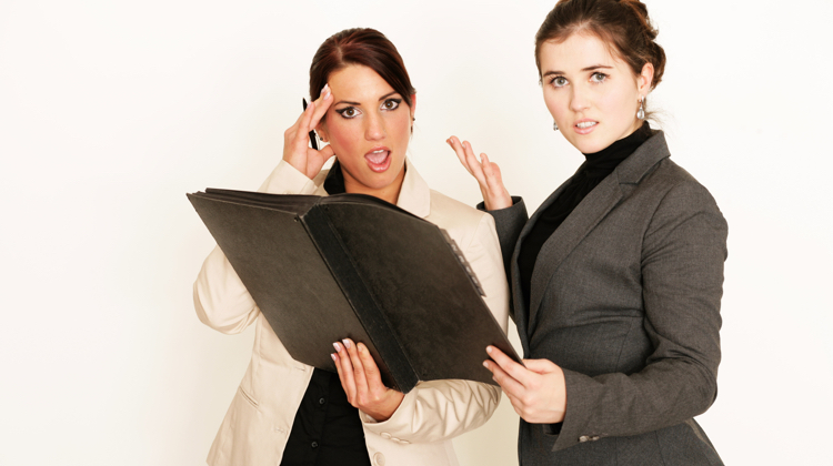 Two women, one in a white blazer and one in a black blazer are looking at a portfolio with shocked expressions, one holding a hand to her head and one holding a hand out as if to say, "what is this?"