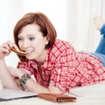 Redheaded woman with short hair and a red-and-white-check shirt lies on her stomach on a bed looking at her laptop and smiling