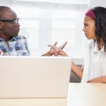 Man and woman face each other at a desk with a laptop open in front of them and point fingers at each other angrily