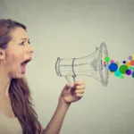 Red-haired woman in white tank top has her mouth open and his holding a pencil-drawn megaphone, which has dots of various colors coming out the speaker side of the megaphone.