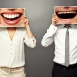Man and woman in white shirts stand against a grey wall and hold up posters with giant mouths that cover their faces.