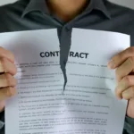 Man in gray, long-sleeved collared shirt holds a paper with the word contract at the top and rips it down the center