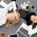 Two women lying on their backs on the floor are surrounded by laptops and iPads while they work