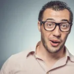 Man in taupe button down shirt and thick black square-rim glasses has his mouth open as if in shock.