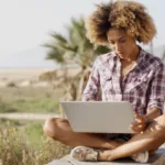 Young woman sits with legs crossed and laptop on lap, with beach scene and palm trees in background.