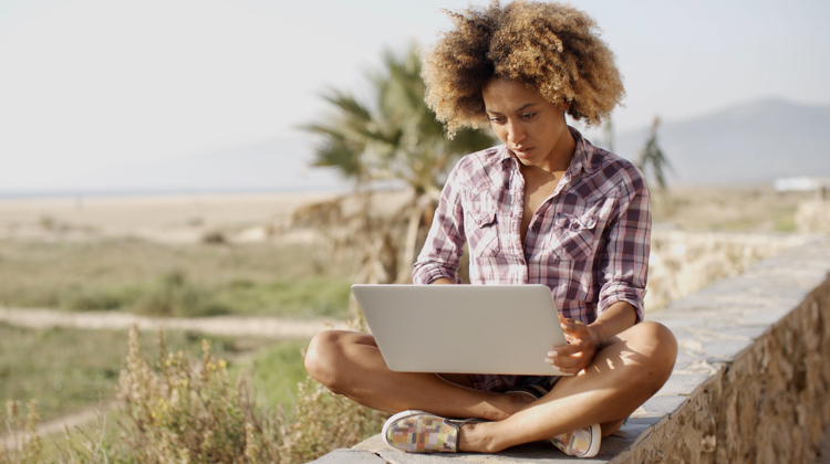 Young woman sits with legs crossed and laptop on lap, with beach scene and palm trees in background.