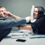 A man and woman in business attire sit across a desk from one another with the man shoving a paper in the woman's face and the woman punching the man's face; HR is not your friend when looking for a job
