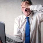 Older man in white dress shirt and blue tie sis in front of a computer with his hand covering his face and his mouth open in surprise.