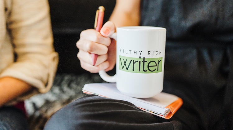 Close-up of hand holding a pen and also gripping white mug sitting on top of a spiral-bound notebook with the words "Filthy Rich Writer" on the mug.