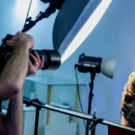 Blonde-haired woman stands against a deep blue background with bright lights and a camera in front of her as she gets her headshot taken.