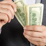 Close-up of hands in front of a suit jacket holding 100 dollar bills as one hand pulls one of the bills away from the stack being held by the other hand.