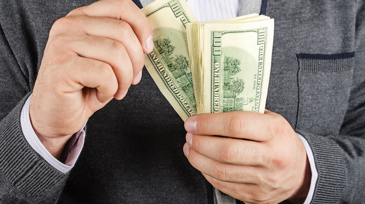 Close-up of hands in front of a suit jacket holding 100 dollar bills as one hand pulls one of the bills away from the stack being held by the other hand.