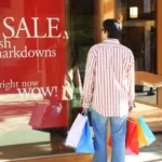 Man in red and white vertical striped shirt and jeans holds five shopping bags of various colors while standing in front of store window that says "sale fresh markdowns."