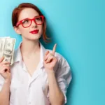 Red-headed woman with hair tied back and red glasses and lips wears a white shirt standing against a teal background holding an array of 100-dollar bills in one hand while pointing over her shoulder with her other hand.