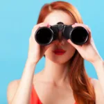 Woman with red hair and orange tank top stands against a blue background holding up binoculars to her face.