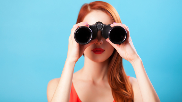 Woman with red hair and orange tank top stands against a blue background holding up binoculars to her face.