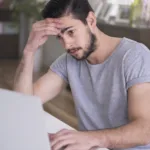 Man rests his head in his left hand and rests his right hand on his laptop with a sad look on face.