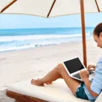 Digital nomad woman works on her laptop from a lounge chair with umbrella that is on the beach in front of the ocean.