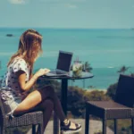 Woman sits at cafe table working on her laptop while overlooking an aqua sea.