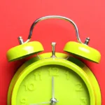 Lime green old-fashioned alarm clock with bells has one hand at the 12 sits against a red background; the 5-second rule is a great way to get down to business.