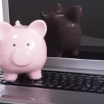 Small pink piggy bank sits on top of the keys of an open laptop.