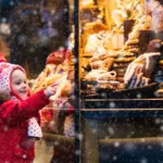 Small child in red snowsuit and hat with tassels reaches for sweets behind a window of a shop as it snows.