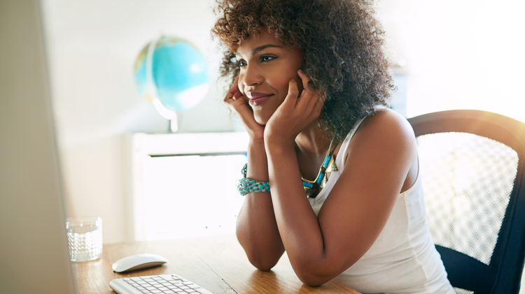 Black woman in white tank top and turquoise bracelet and necklace smiles while holding her face in her hands as she looks at a monitor with a keyboard in front of her and globe out of focus in the background.