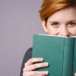 Woman with short red hair and grinning eyes holds a teal book up to hide her face.