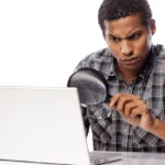 Black man in flannel shirt with rolled up sleeves raises an eyebrow as he looks through a magnifying glass at a laptop.