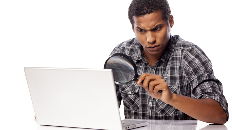 Black man in flannel shirt with rolled up sleeves raises an eyebrow as he looks through a magnifying glass at a laptop.