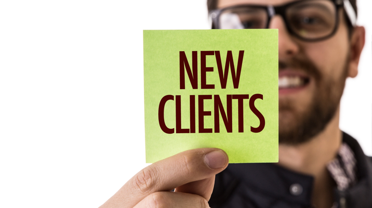 Man with glasses and beard holds a lime green sticky note in front of his face that says "new clients" in all capital block letters.