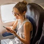 Woman in an airplane seat by window is smiling while looking at phone.