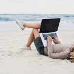 Person on beach in jean shorts, taupe top, and a straw hat lays with their canvas duffle bag as a pillow with their laptop on their knees.