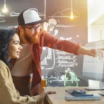 Bearded man in glasses and trucker hat leans over desk and points at screen collaborating with the woman who is sitting at the desk.