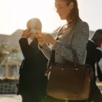 Woman in wool blazer and brown leather bag looks at her phone while people in business attire pass around her as the sun starts to set.