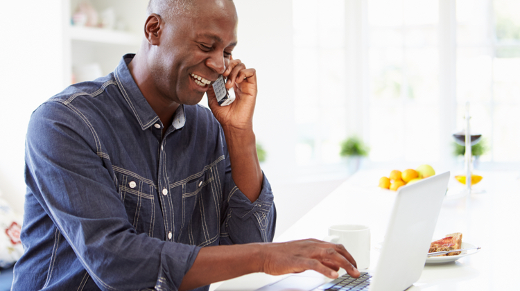 Bald Black man in a denim top laughs while talking on the phone and typing on his laptop.