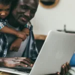 Smiling man wearing a flannel shirt sits in front of his laptop with a young boy hugging him from behind